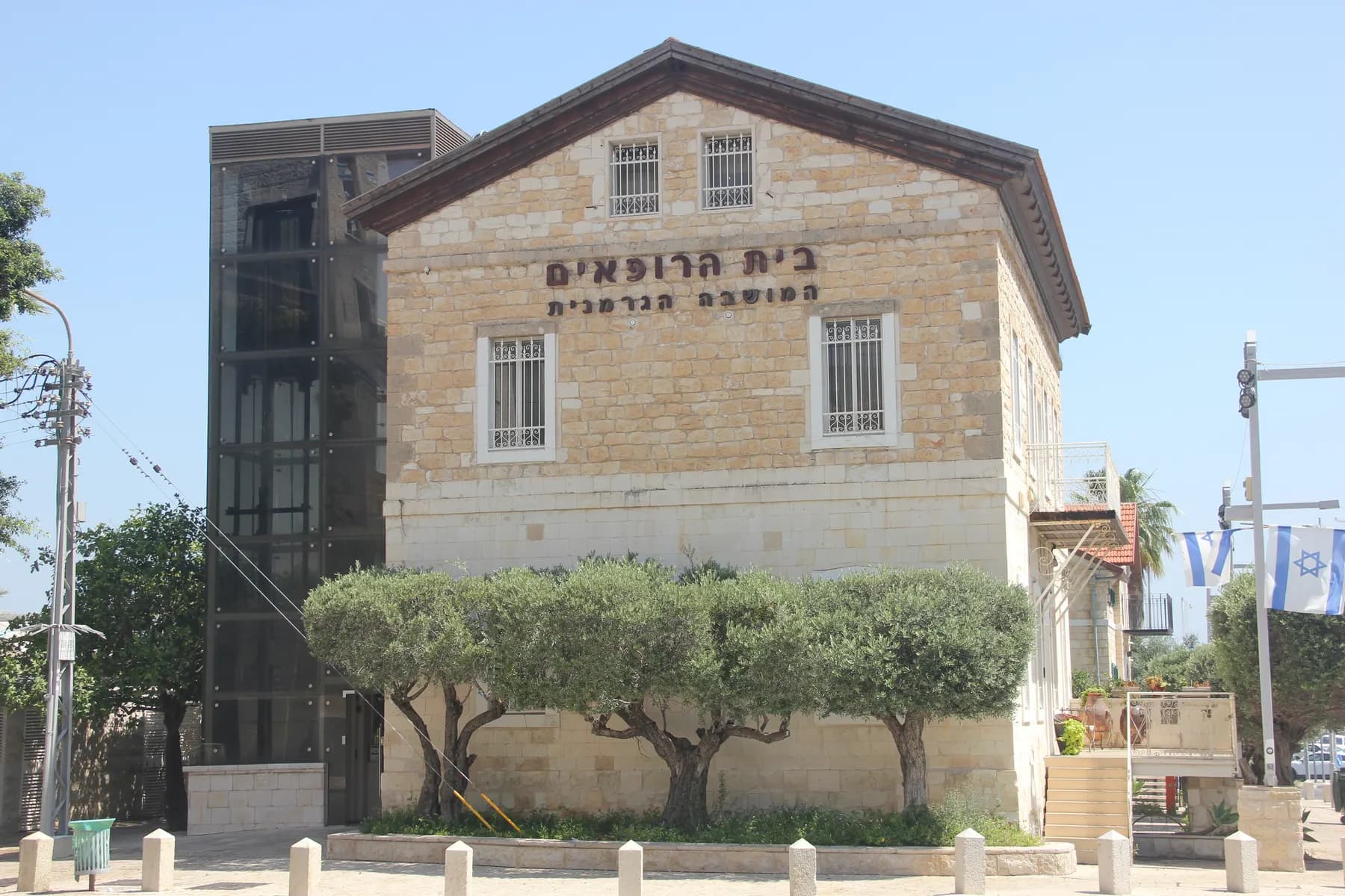 A two-storey Templer house of pale dressed stone in Haifa's German Colony, its facade carrying a Hebrew inscription, with clipped olive trees along the pavement in front