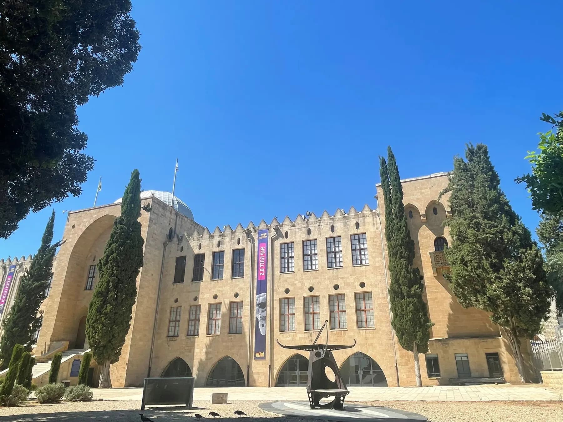 The crenellated sandstone front of the historic Technion building in Haifa, its rows of arched windows framed by tall cypress trees under a clear blue sky