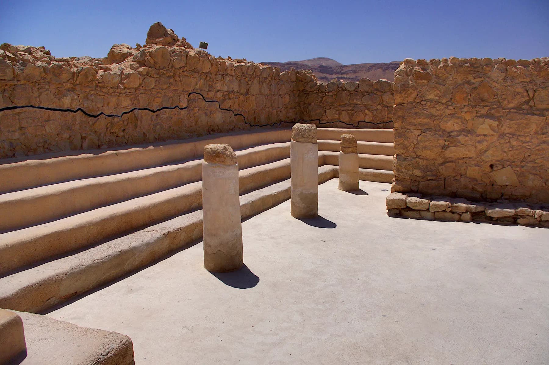 Tiered stone benches and short column drums inside the excavated synagogue on the summit of Masada, with desert hills beyond the wall