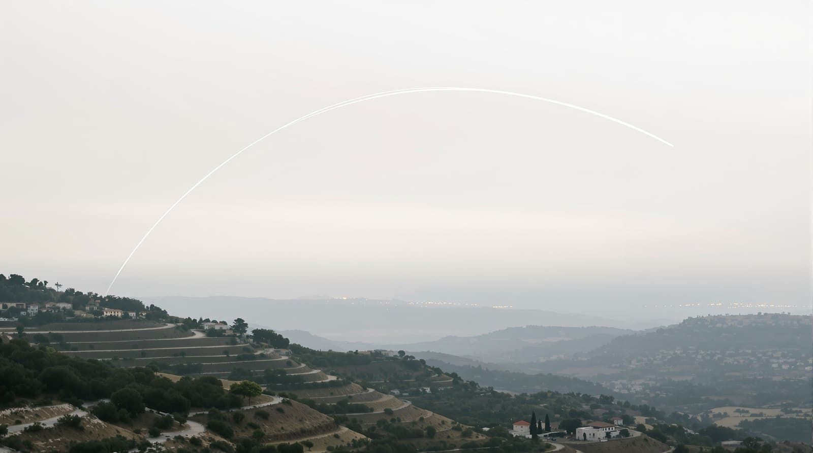 Illustration of a thin white contrail curving over the hazy hills of southern Lebanon, seen from a ridge of olive terraces at dawn.