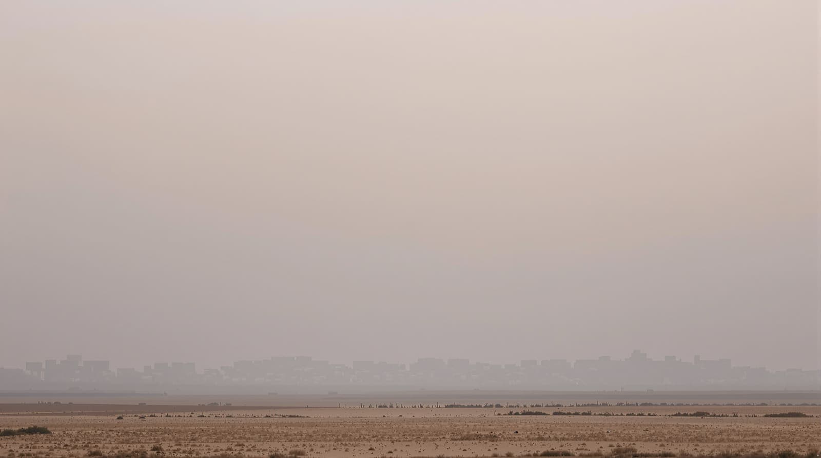 Illustration of the hazy low skyline of Khan Yunis in southern Gaza seen from distant sandy farmland at dawn.