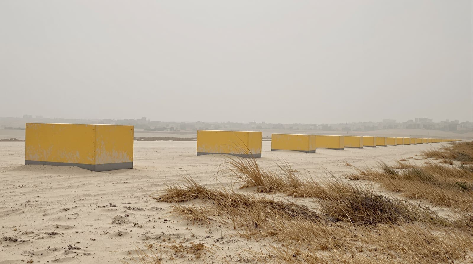 Illustration of a spaced row of yellow concrete blocks set across sandy ground in northern Gaza, seen from a low rise under an overcast sky.
