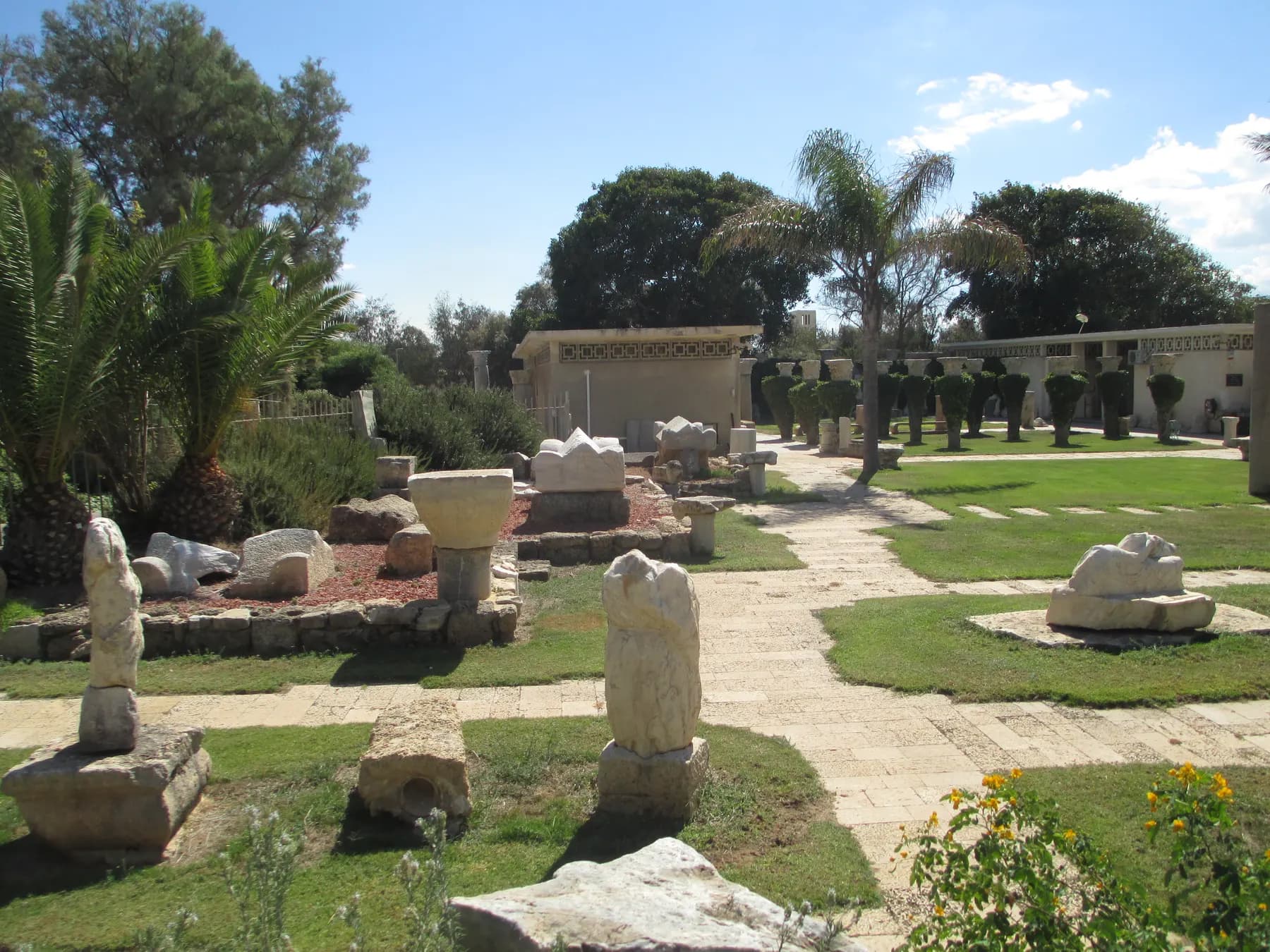 Roman column capitals, carved marble blocks and a broken stele set out on the lawn and gravel paths of the museum garden, with the low colonnaded museum building behind