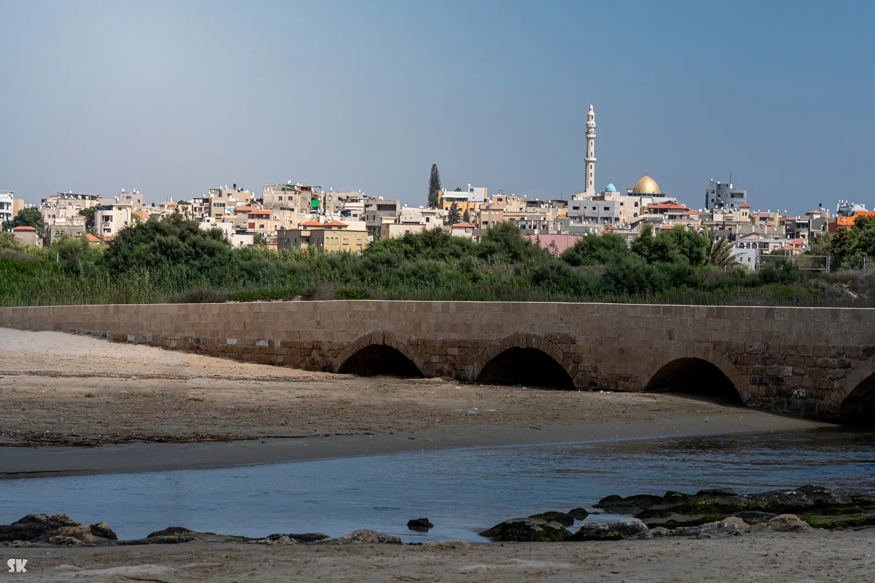 An old stone bridge on three arches spanning a sandy stream mouth, with the houses, minaret and golden dome of Jisr az-Zarqa on the rise behind
