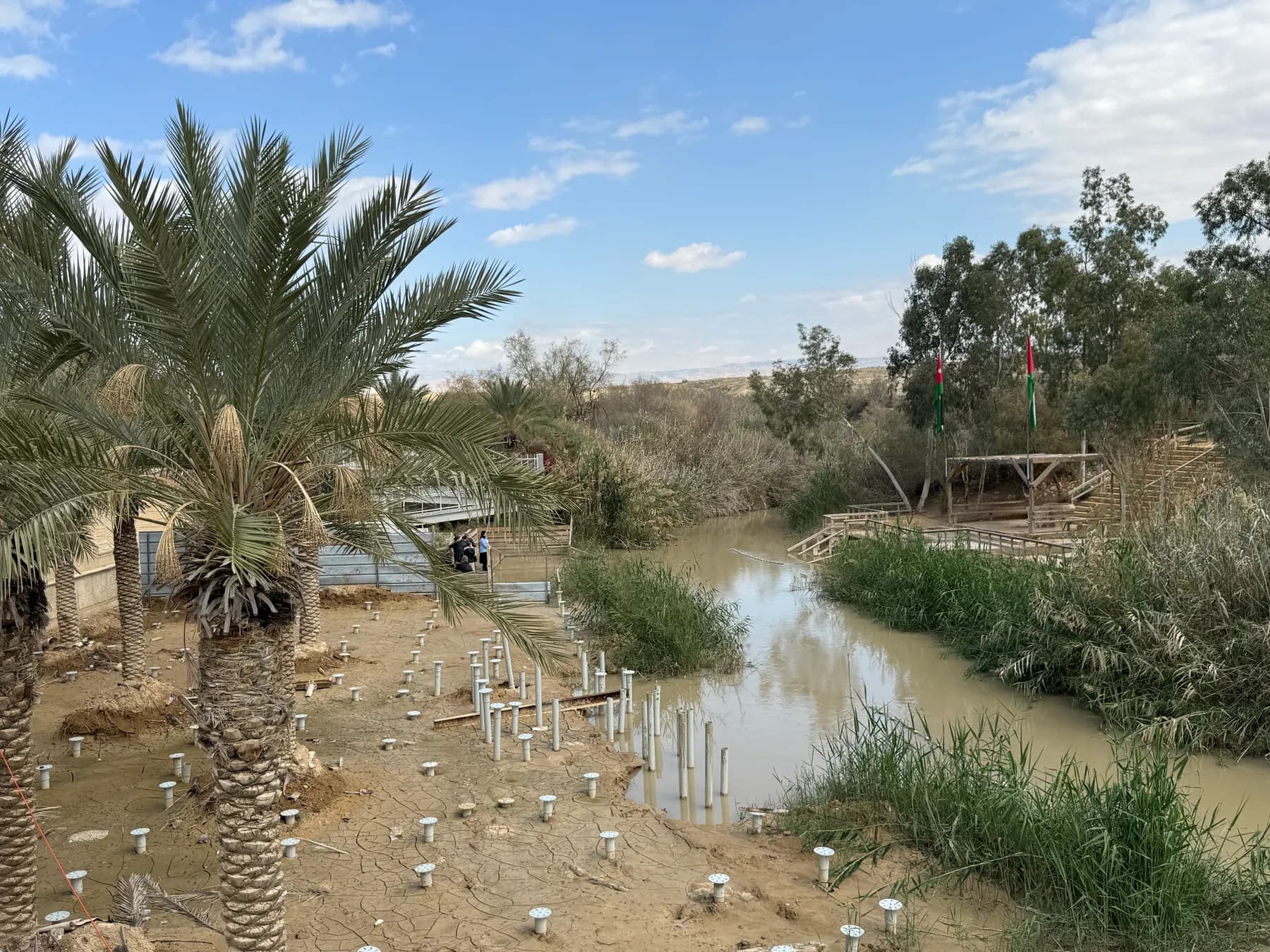 Date palms and a railed walkway on the near bank of the Jordan River at Qasr al-Yahud, with a flagged wooden platform and ramp on the bank opposite