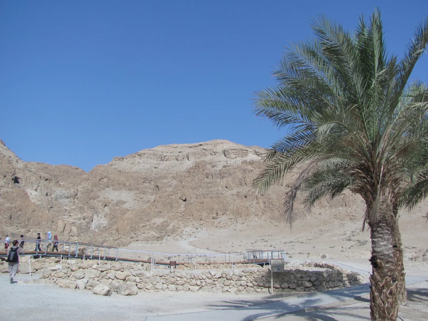 Visitors on the raised metal walkway above the low excavated stone walls at Qumran, with the desert cliffs rising behind