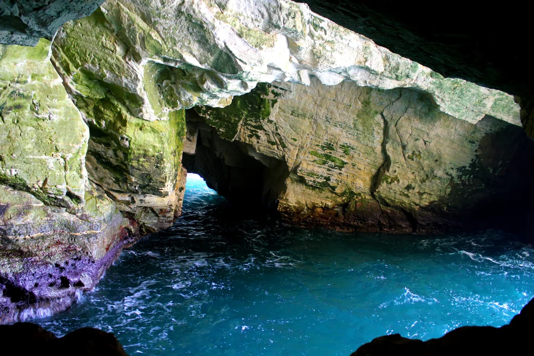 Inside a sea grotto at Rosh Hanikra, pale chalk walls streaked with green arching over turquoise water breaking white against the rock