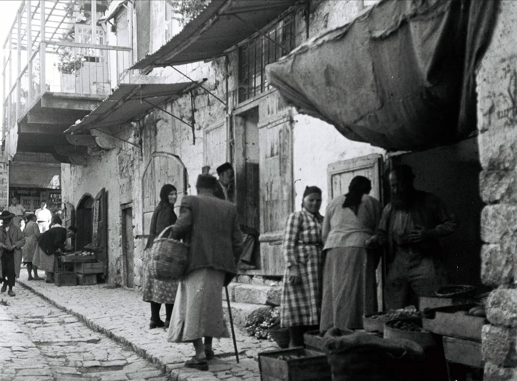 A black-and-white view along a stone-paved lane in Safed's old city, cloth awnings shading a row of stone shopfronts with people standing among the stalls