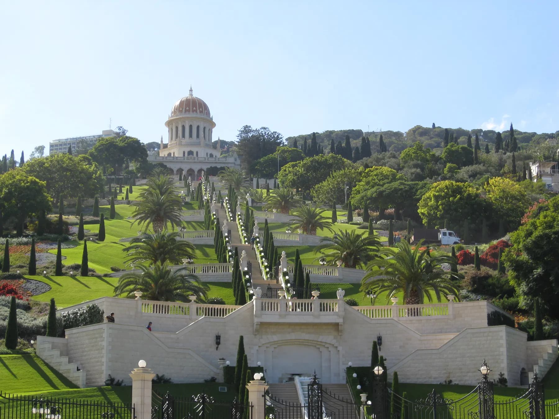 White terraces and a central stairway climbing a green hillside between cypresses and palms to a gold-domed shrine