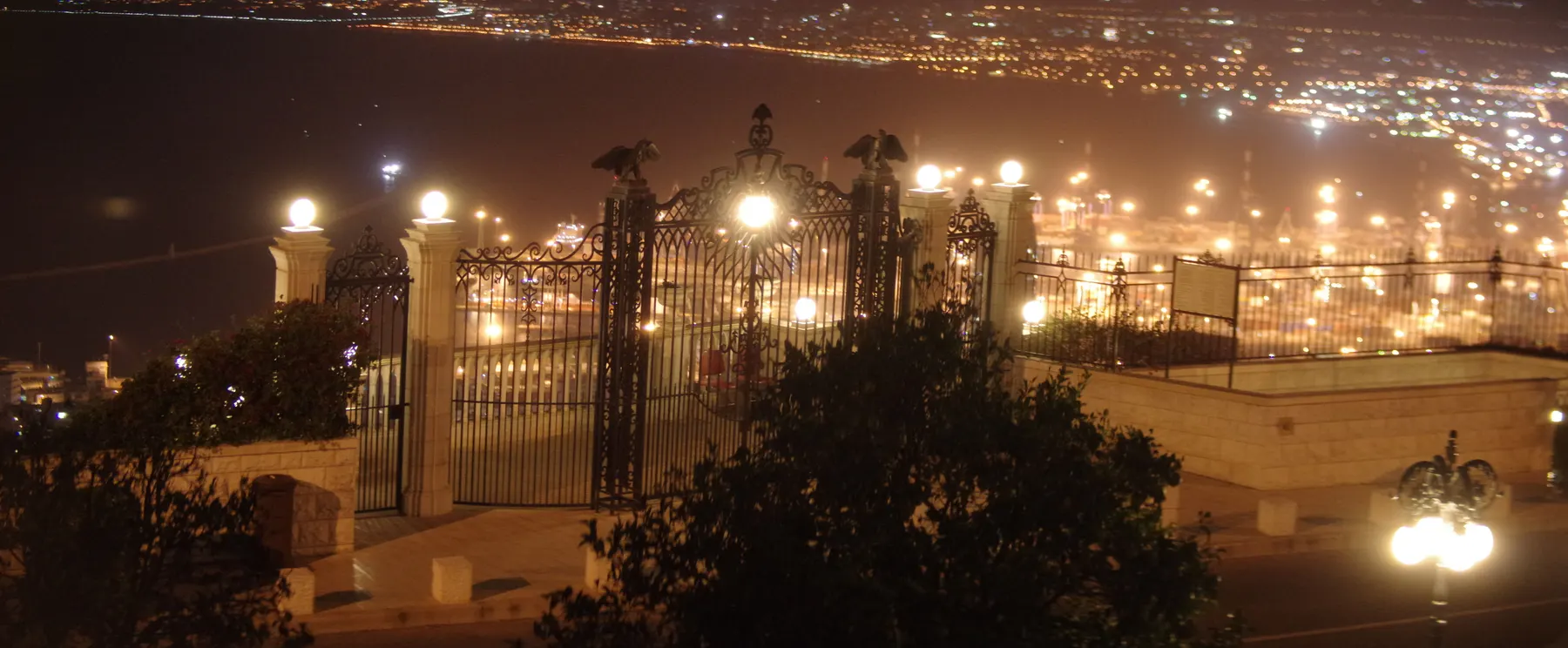 An ornamental iron gate and cliff-edge railing lit at night on the Carmel ridge, with the lights of Haifa Bay curving away below