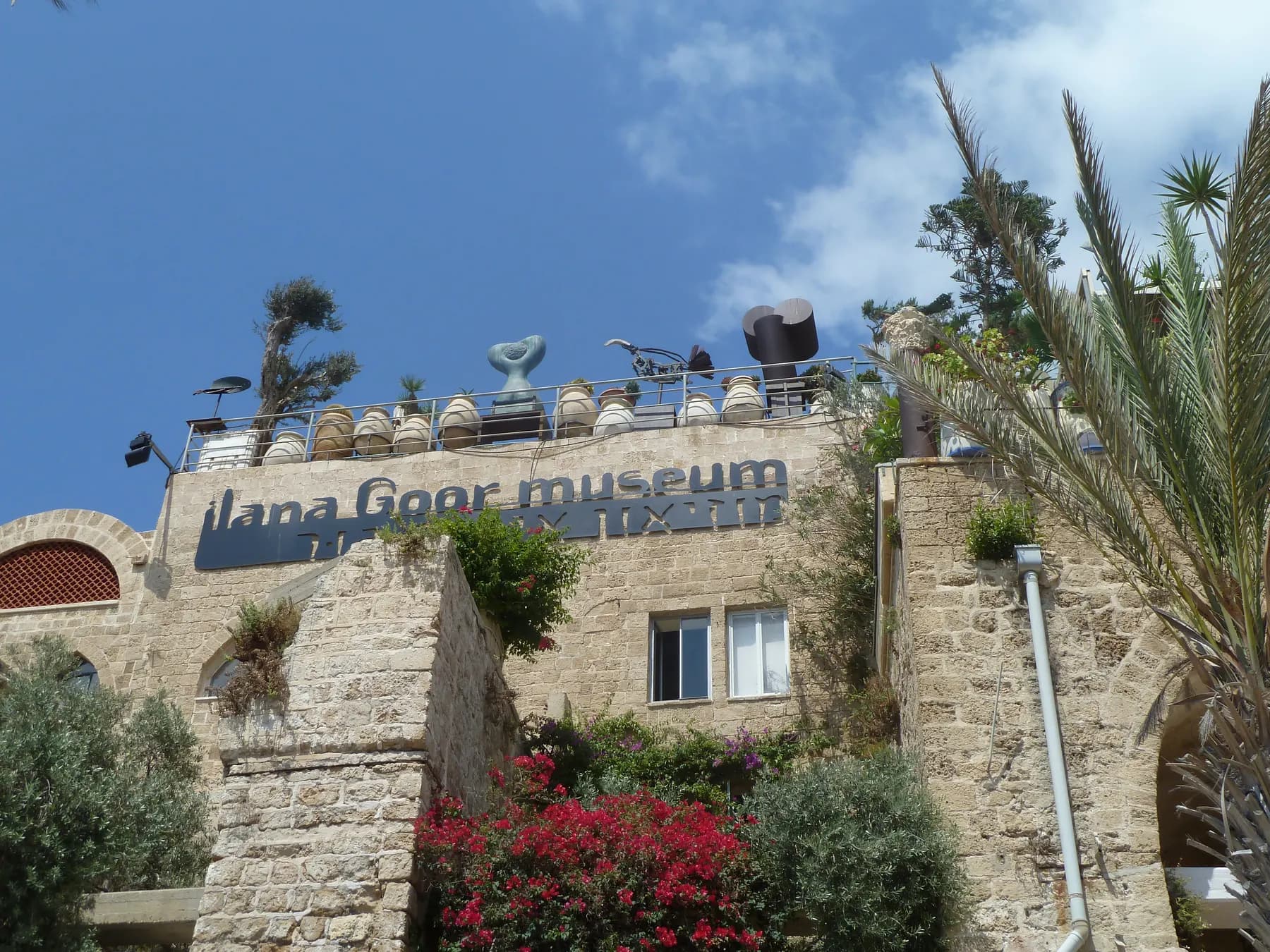 The stone facade of the Ilana Goor Museum in Old Jaffa, its painted name below a roof terrace lined with sculptures and rows of pottery jars, bougainvillea flowering against the wall