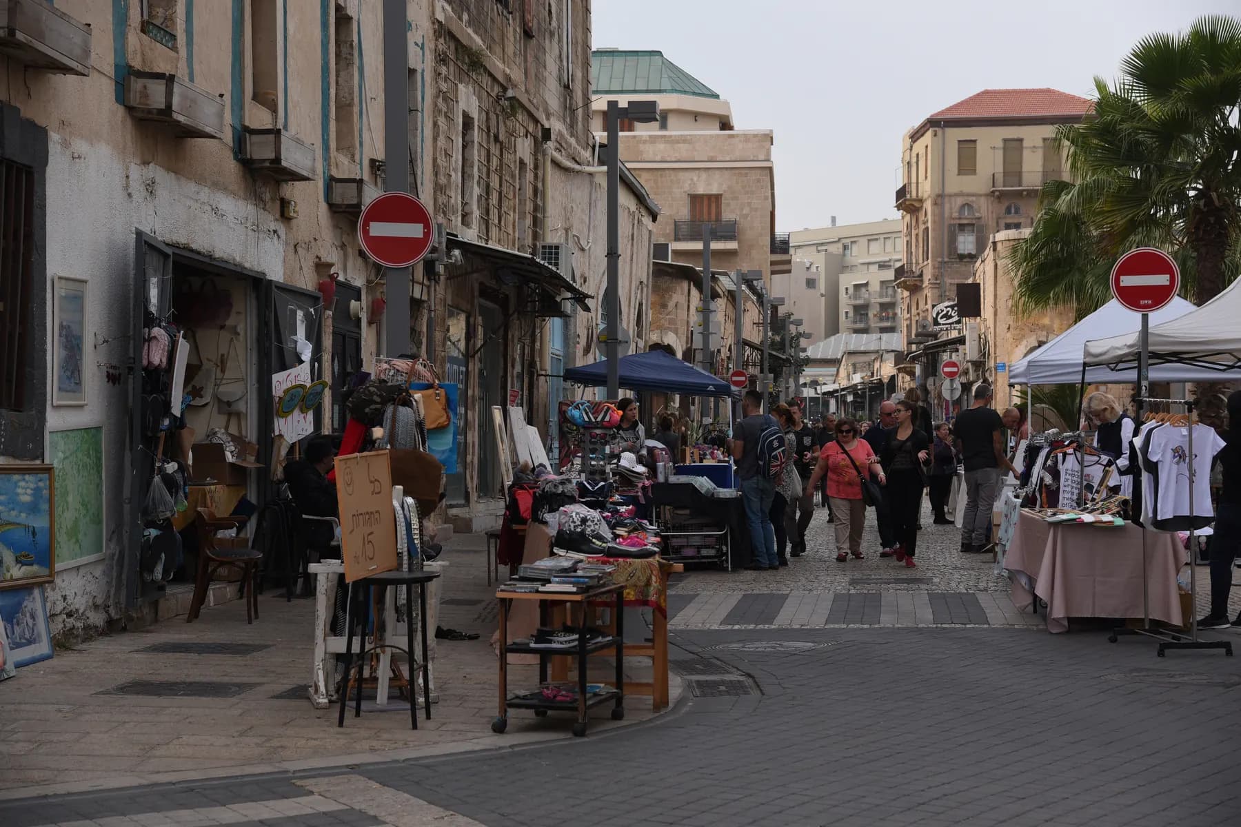 A narrow Jaffa market lane lined with stalls of framed pictures, second hand furniture and clothing racks, with shoppers walking between them