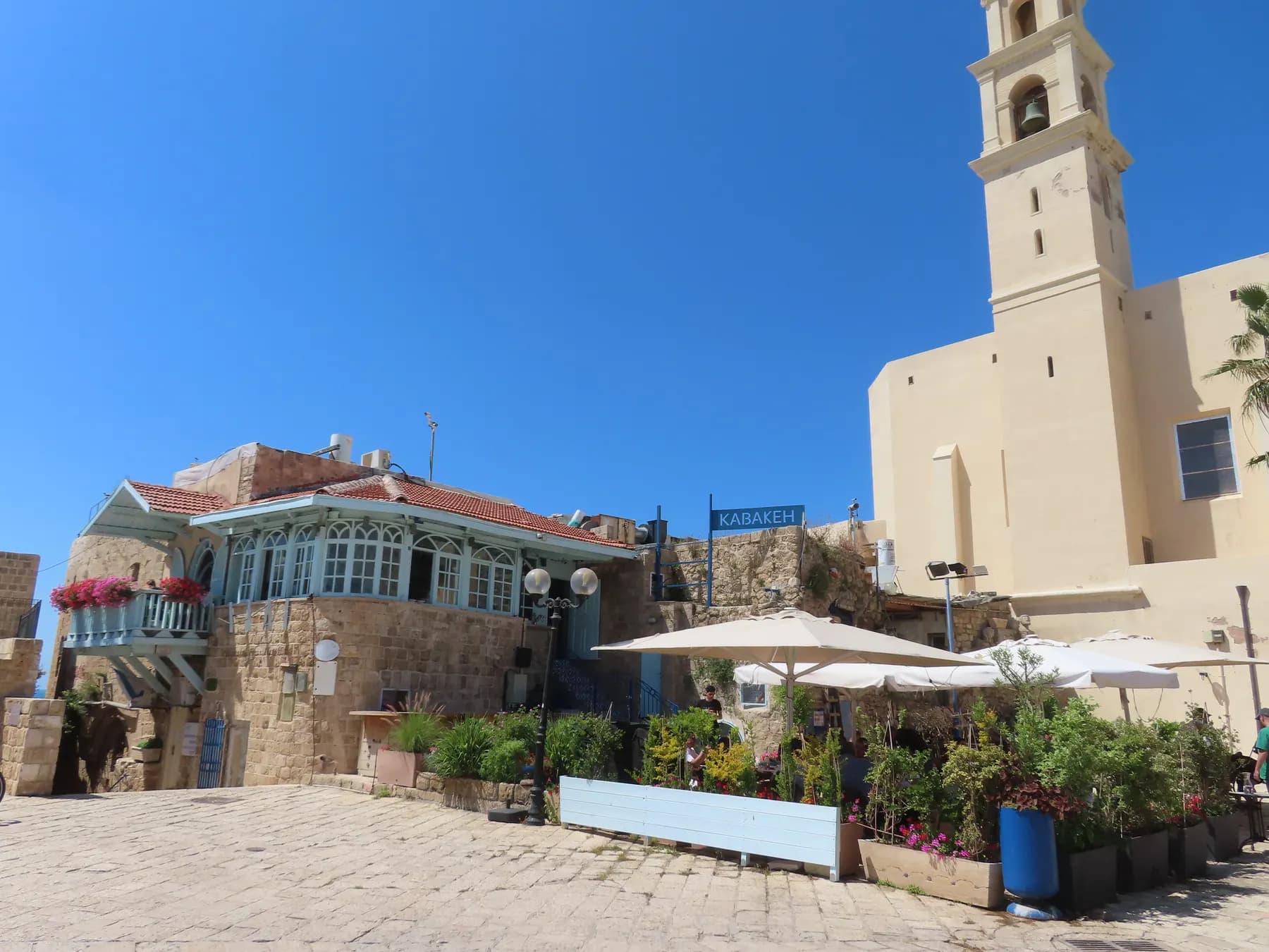 The paved square in Old Jaffa with the bell tower and cream stone wall of St Peter's Church rising on the right, above cafe umbrellas, planters and a blue and white wooden veranda house