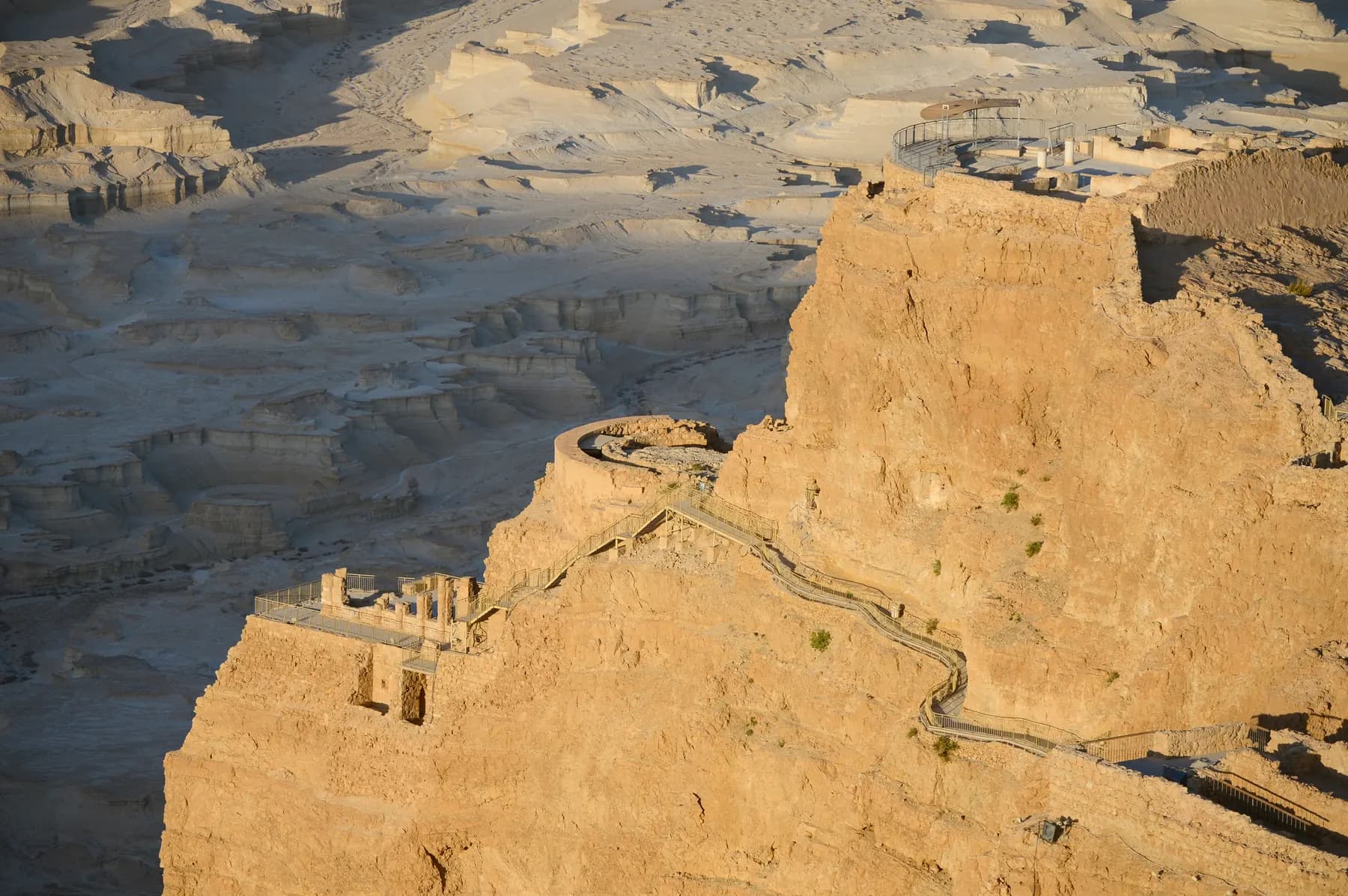The tiered terraces of Herod's palace built into the northern cliff of Masada in low golden light, a round colonnaded terrace above a lower terrace of standing columns with a stepped path linking them