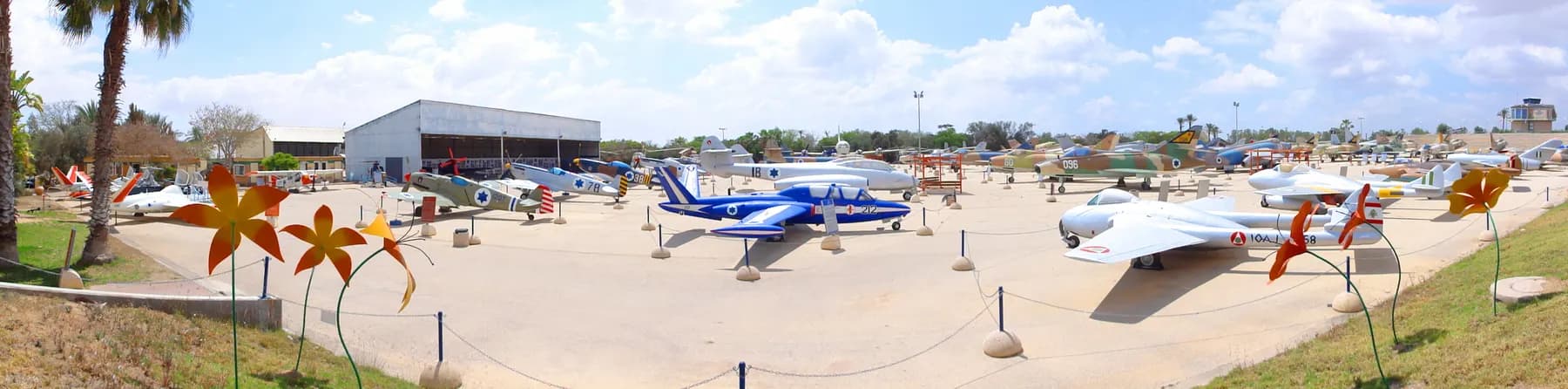 Rows of historic Israeli Air Force aircraft parked on a concrete apron in front of an open hangar under a bright cloudy sky