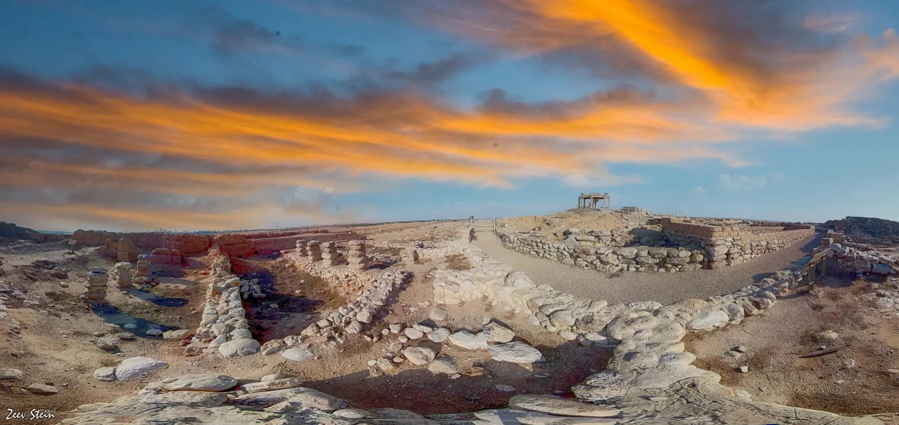 Excavated stone walls and lanes covering the flat top of the mound at Tel Be'er Sheva, under a streaked orange sunset sky