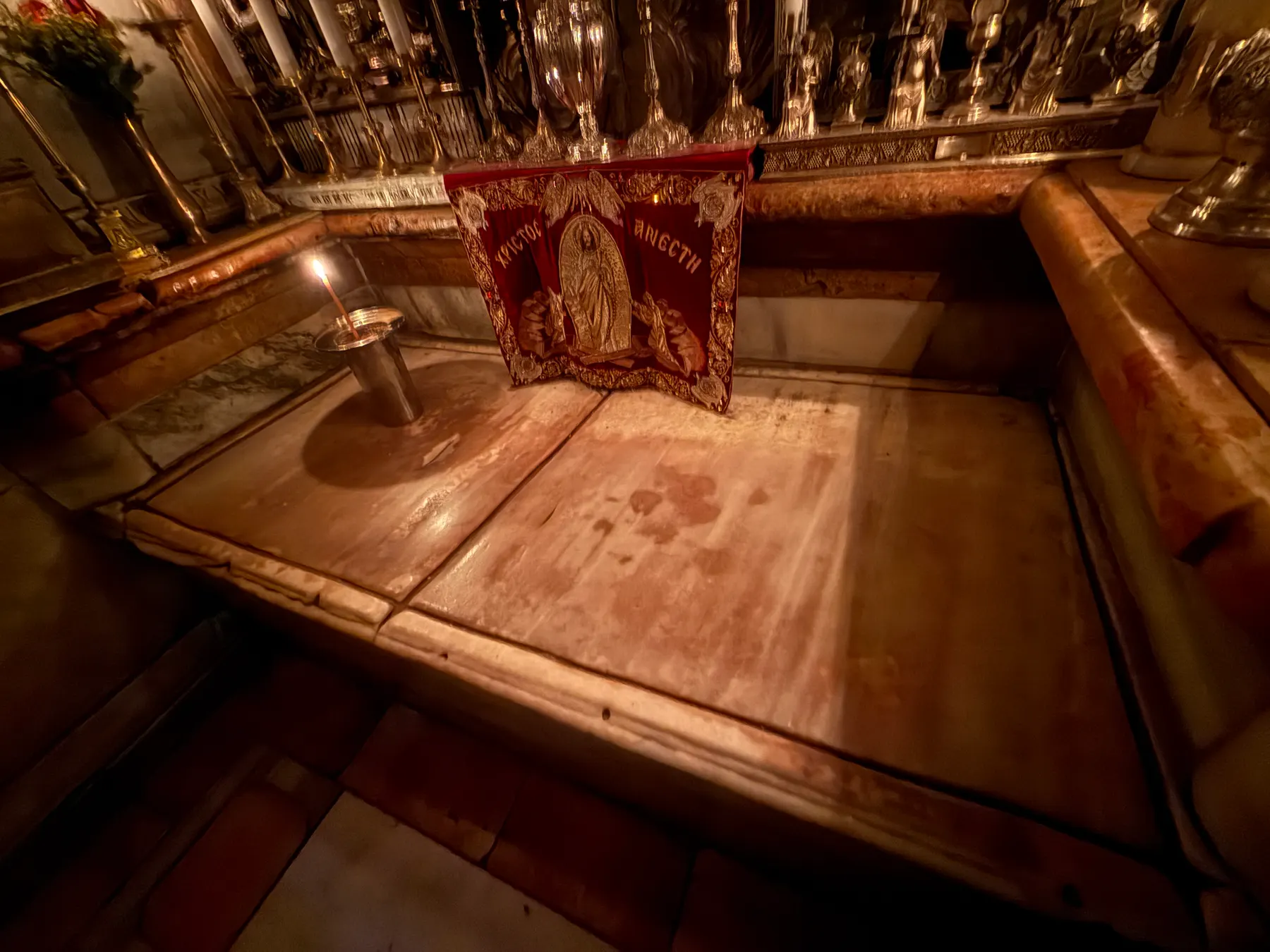 The marble slab of the tomb inside the Aedicule, lit by a single candle beside a red embroidered banner