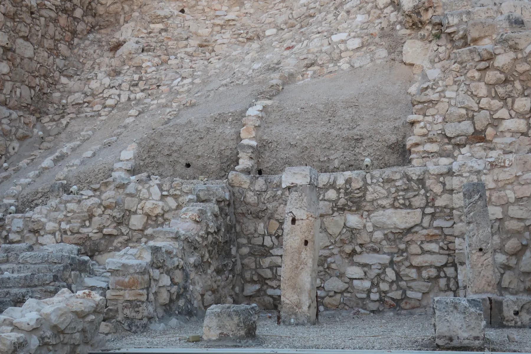 Excavated stone house walls with two upright monolithic pillars, built against the steep earth and rubble slope of the ridge