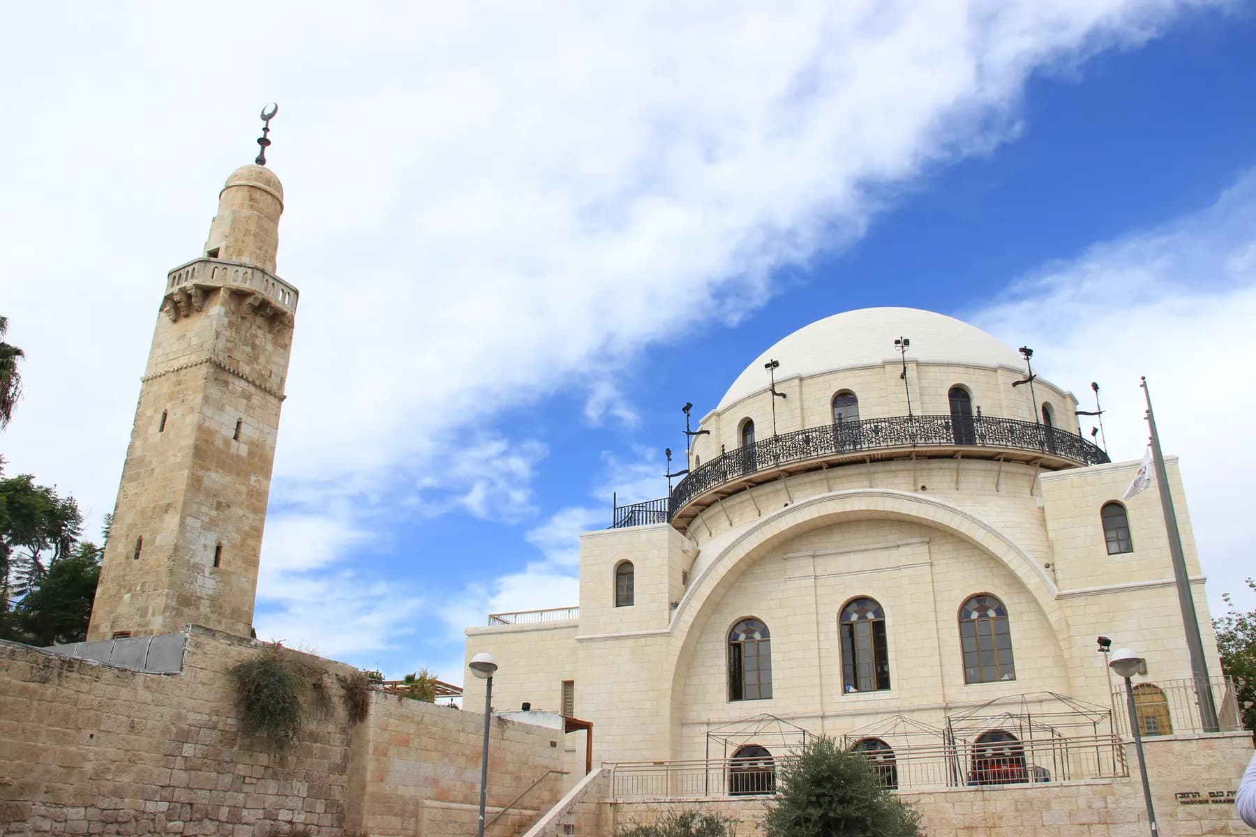 The white dome and tall arched stone facade of the Hurva Synagogue, with a slender minaret rising beside it against a cloudy sky
