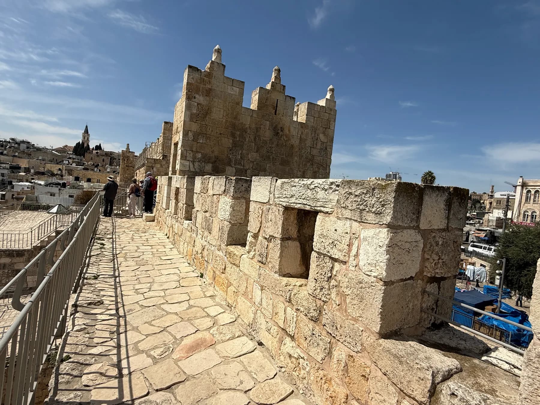 A stone-paved walkway running along the top of the Old City wall between crenellated battlements toward a square gate tower, with a few walkers further along