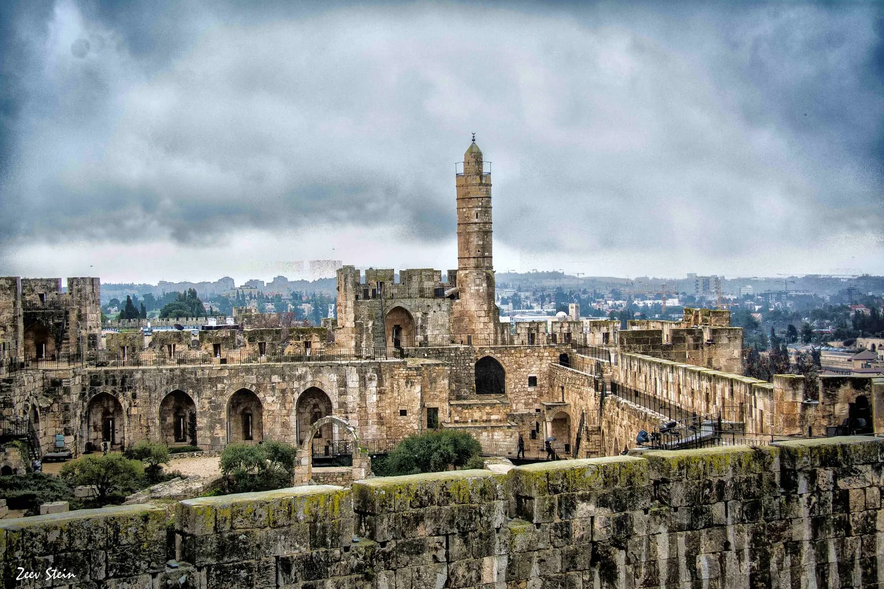 The Tower of David citadel in Jerusalem seen from its ramparts, its tall stone minaret rising above the arched courtyard under a heavy grey sky