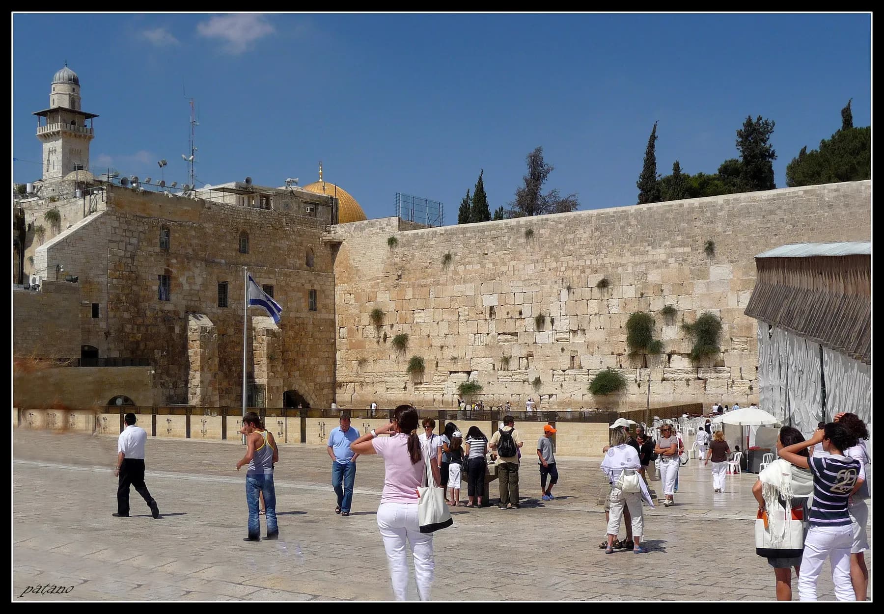 The Western Wall's tiers of pale stone blocks above the open plaza in Jerusalem, with an Israeli flag, a minaret and the gold Dome of the Rock behind