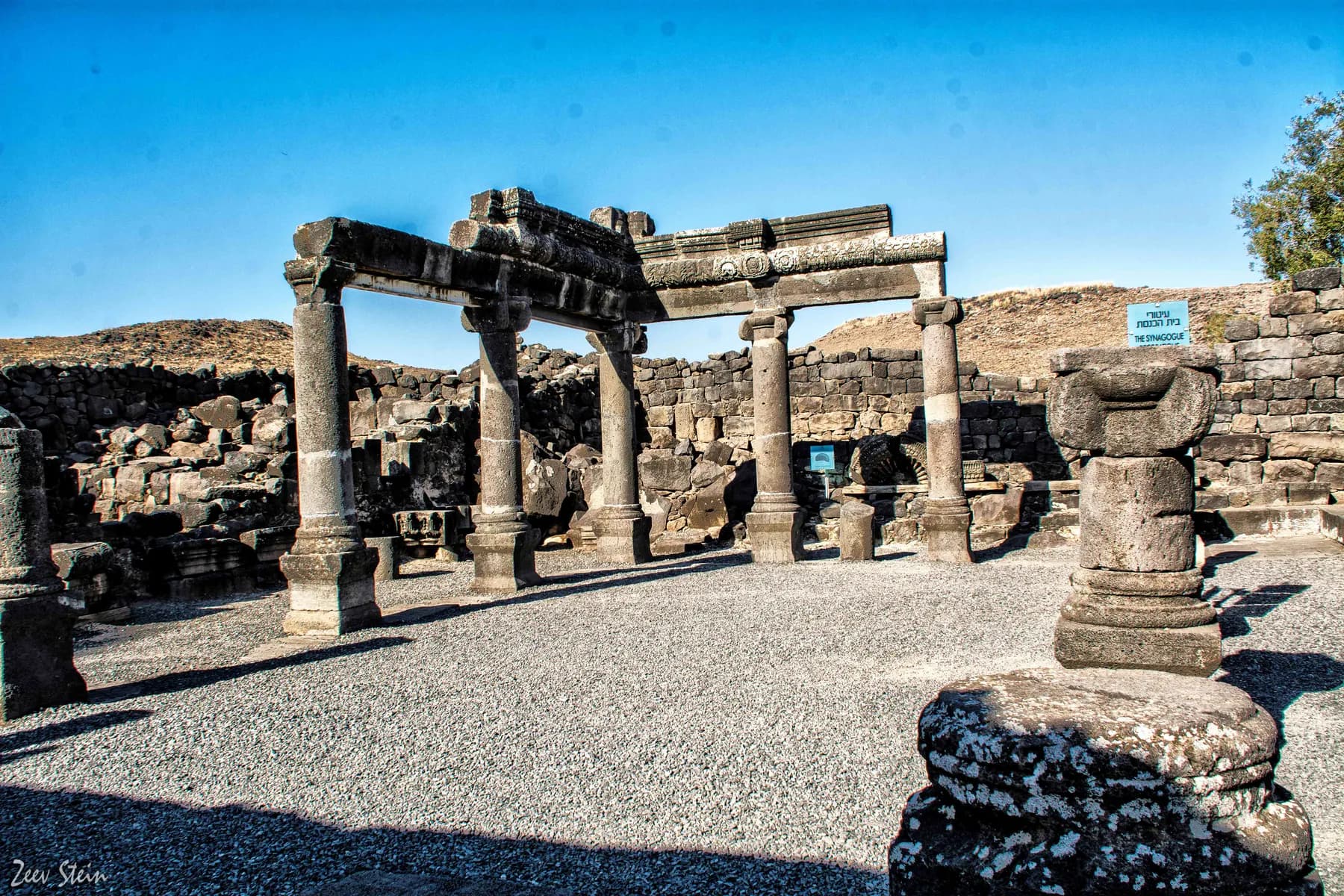Reconstructed dark basalt columns carrying a carved lintel at the Korazim synagogue, with low ruined walls and bare hills behind