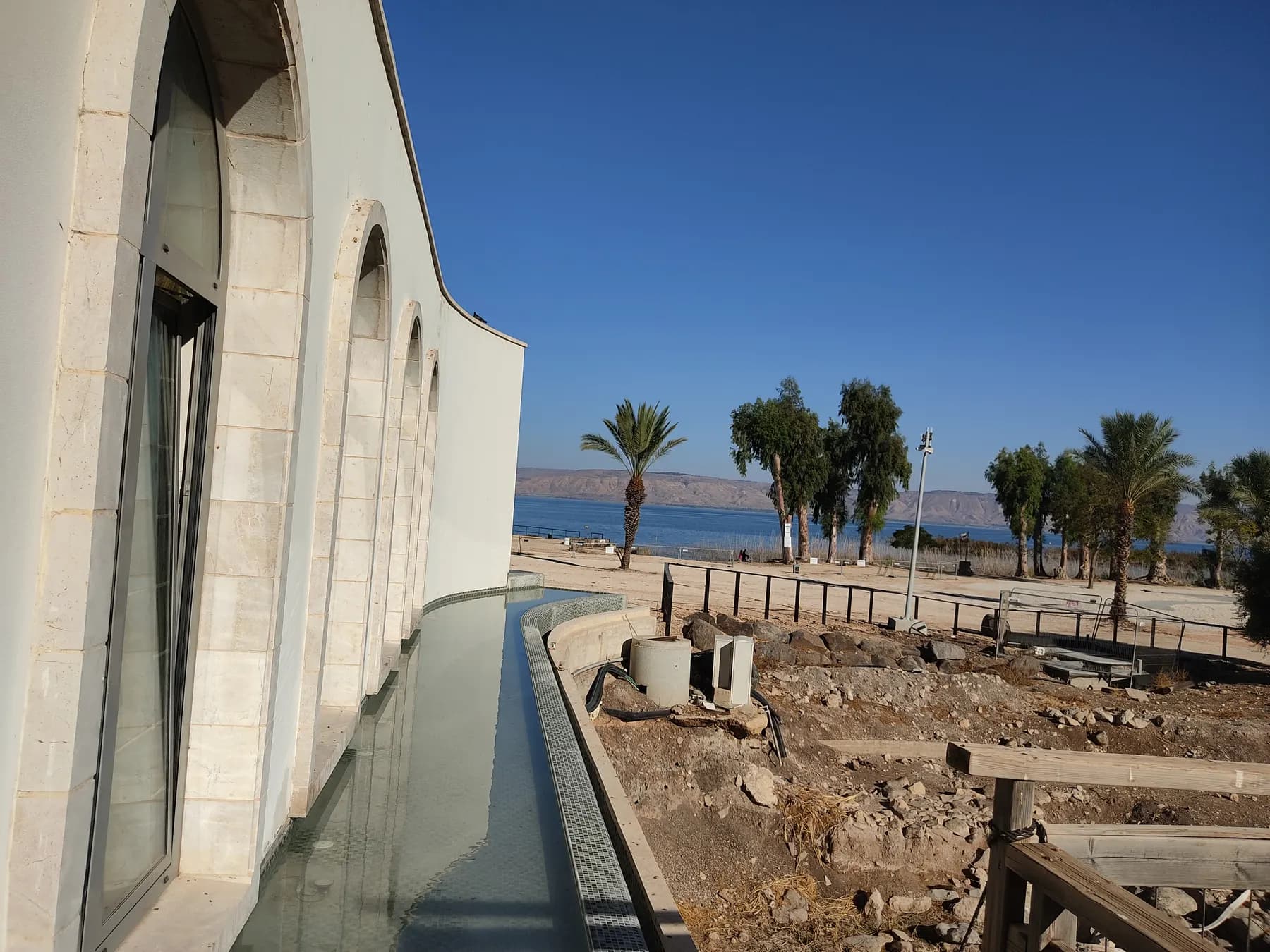 The curved white facade and tall arched windows of the Duc in Altum chapel above a shallow reflecting channel, with palms and the Sea of Galilee beyond
