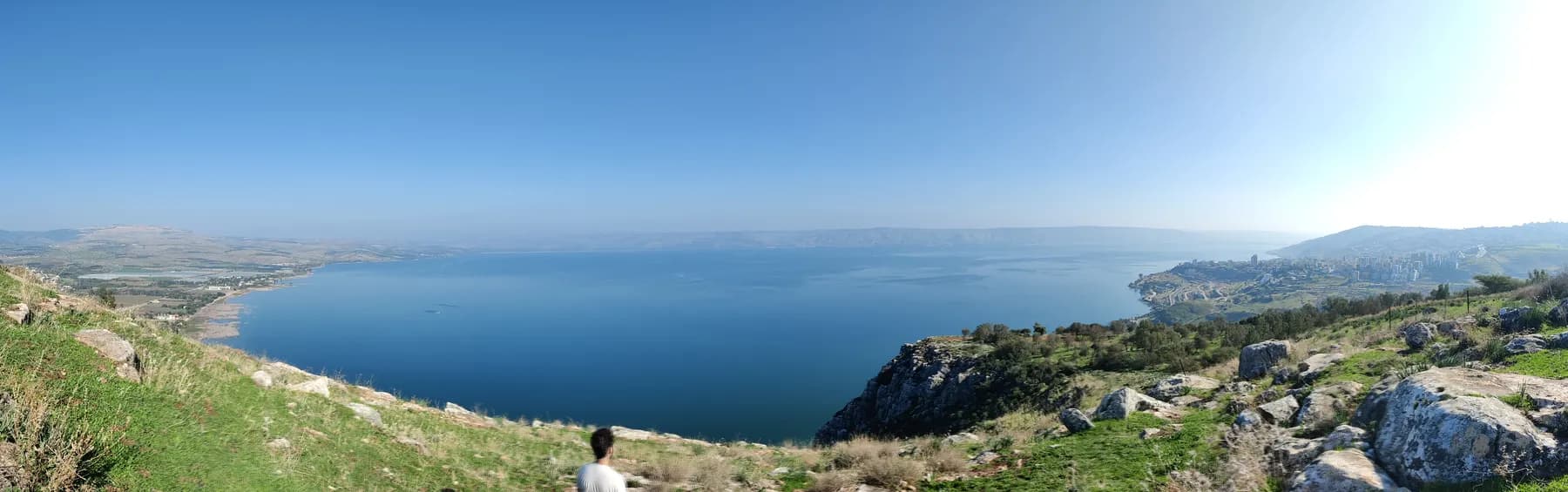 A panoramic view east over the blue expanse of the Sea of Galilee from the grassy clifftop of Mount Arbel, with a rocky spur falling away towards the shore
