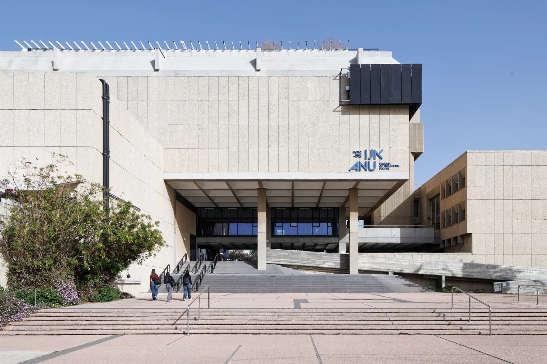 The concrete facade of ANU, the Museum of the Jewish People, its blue ANU sign set above a recessed entrance portico reached by broad steps