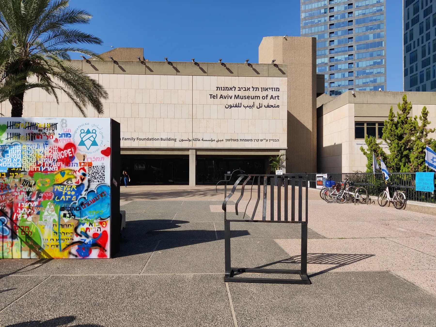 The stone facade of the Tel Aviv Museum of Art with its name in Hebrew, English and Arabic above the recessed entrance, a sticker-covered panel and bicycle racks on the plaza in front