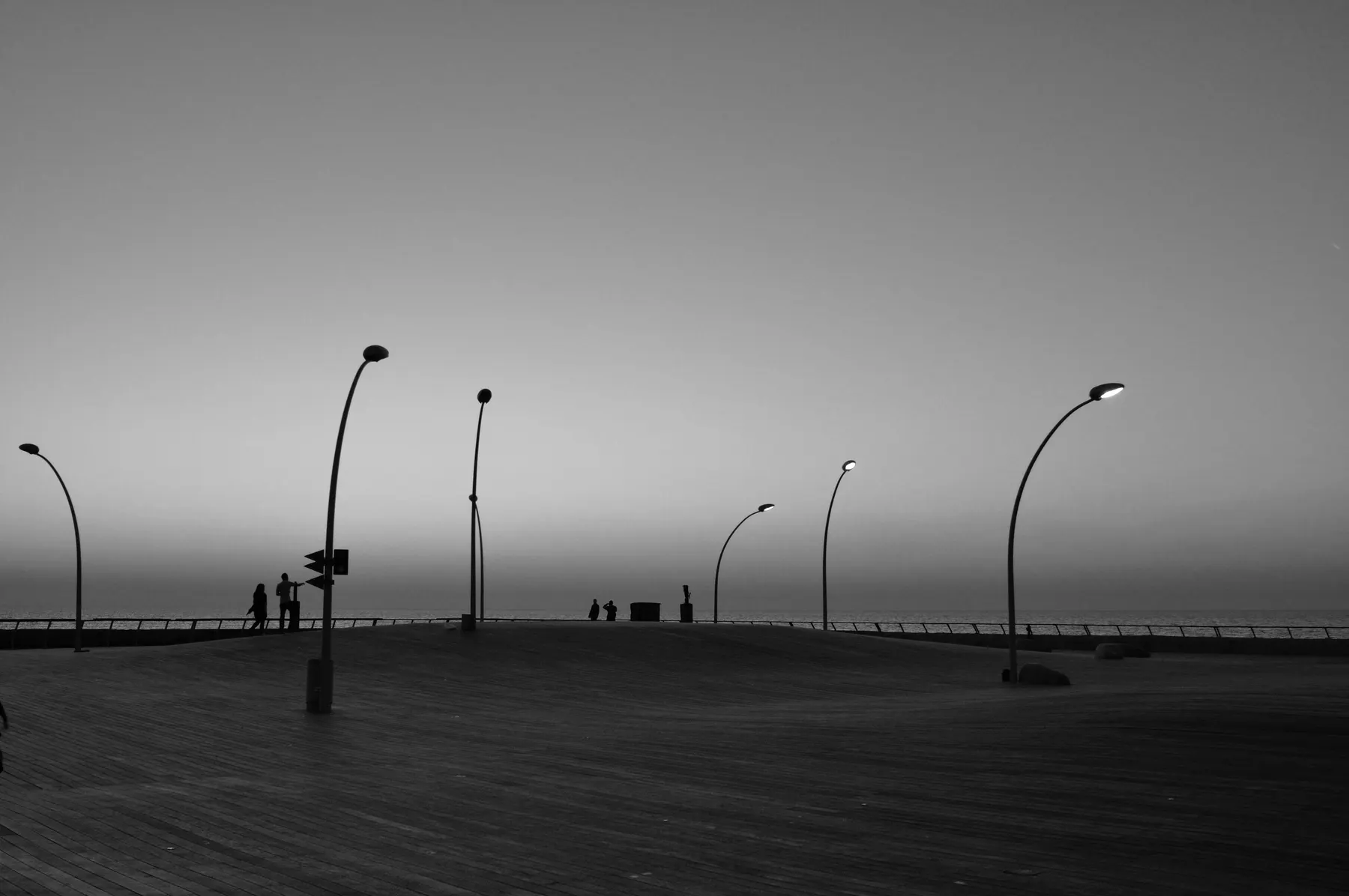 The broad timber deck of the port at dusk, curved lamp posts rising from it and the flat sea horizon beyond, in black and white