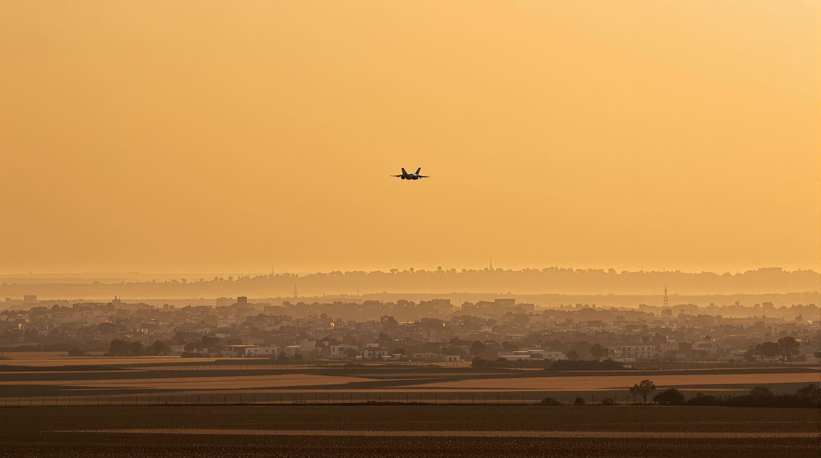 Illustration of a fighter jet silhouetted at dusk above a hazy Gaza skyline, with a kibbutz perimeter fence in the foreground.